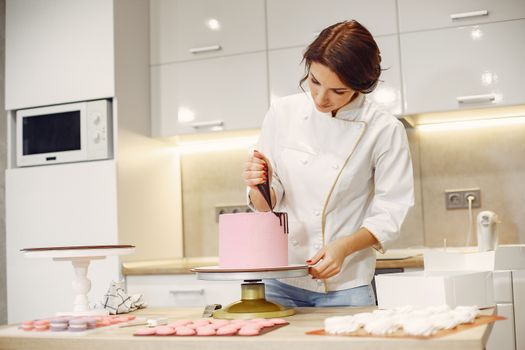 Pâtissière décorant un gâteau rose dans une cuisine moderne, entourée de macarons et de meringues.