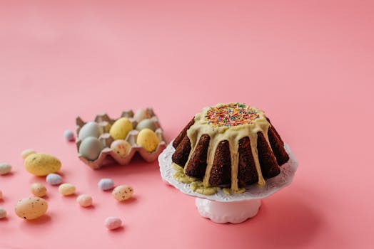 Gâteau de Pâques décoré de glaçage jaune et de vermicelles, entouré d'œufs en chocolat sur fond rose.
