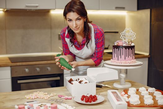 Pâtissière en train de décorer un gâteau rose avec des macarons et des douceurs sur la table de cuisine.