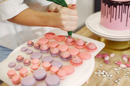 Pâtissière en train de décorer des macarons roses et violets sur un plateau, avec un gâteau en arrière-plan.